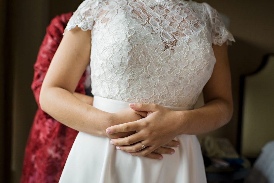 A bride holds her hands while standing in her lace wedding dress.